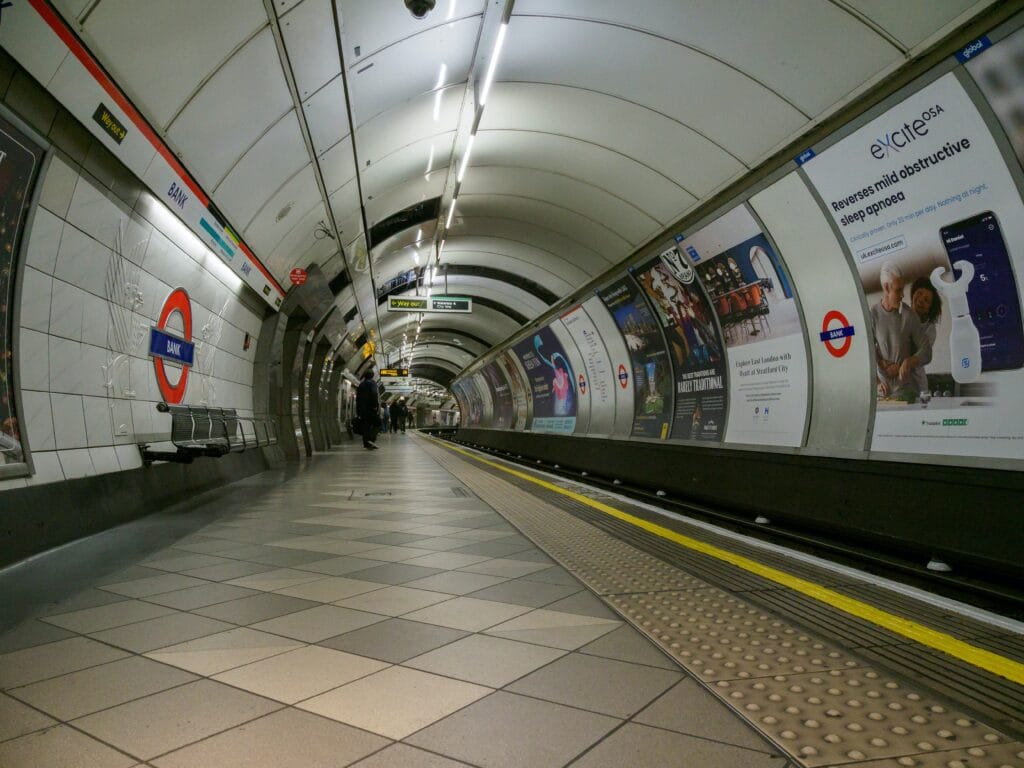 A platform at an Underground subway station in London. Tactile paving can be seen and multiple advertisement posters on the walls.