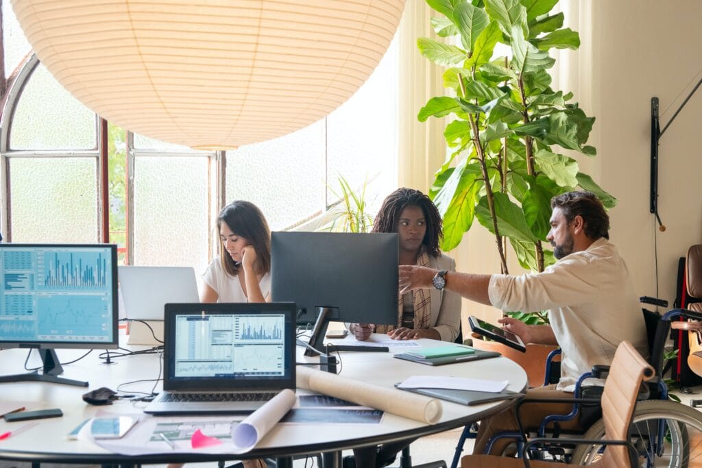 A mixed group of work colleagues discuss work around an office table. One of them, a male wheelchair user, points to his female colleagues monitor to point something out.