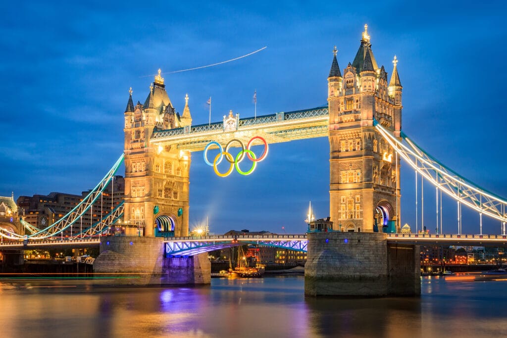 A panoramic photograph of a lit-up Tower Bridge in London during the evening. The Olympics logo is being hung from the top centre of the bridge.