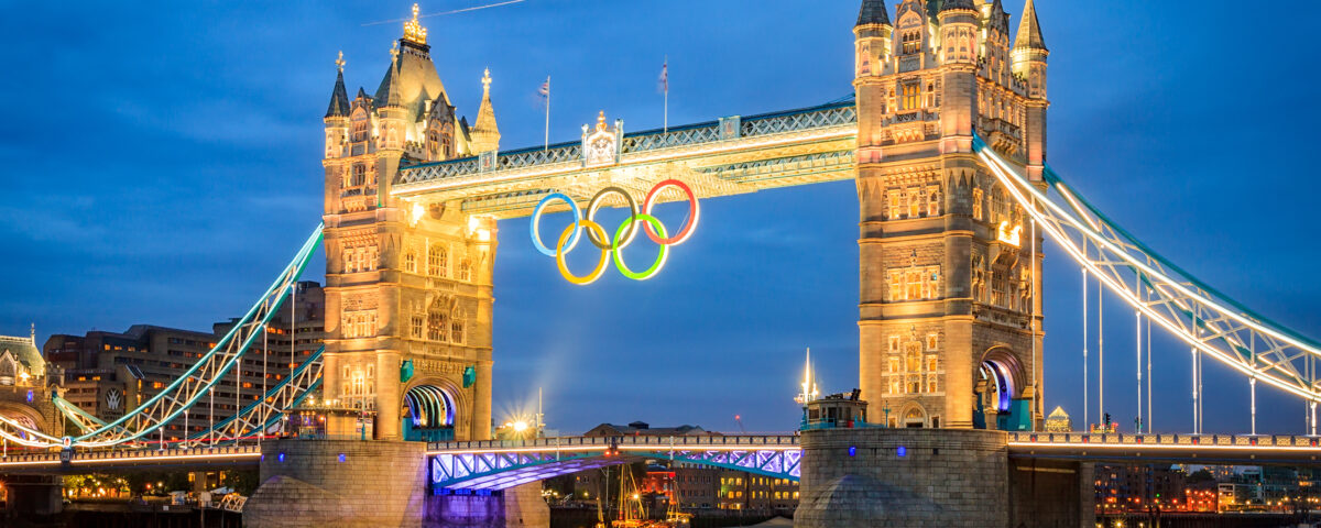 A panoramic photograph of a lit-up Tower Bridge in London during the evening. The Olympics logo is being hung from the top centre of the bridge.