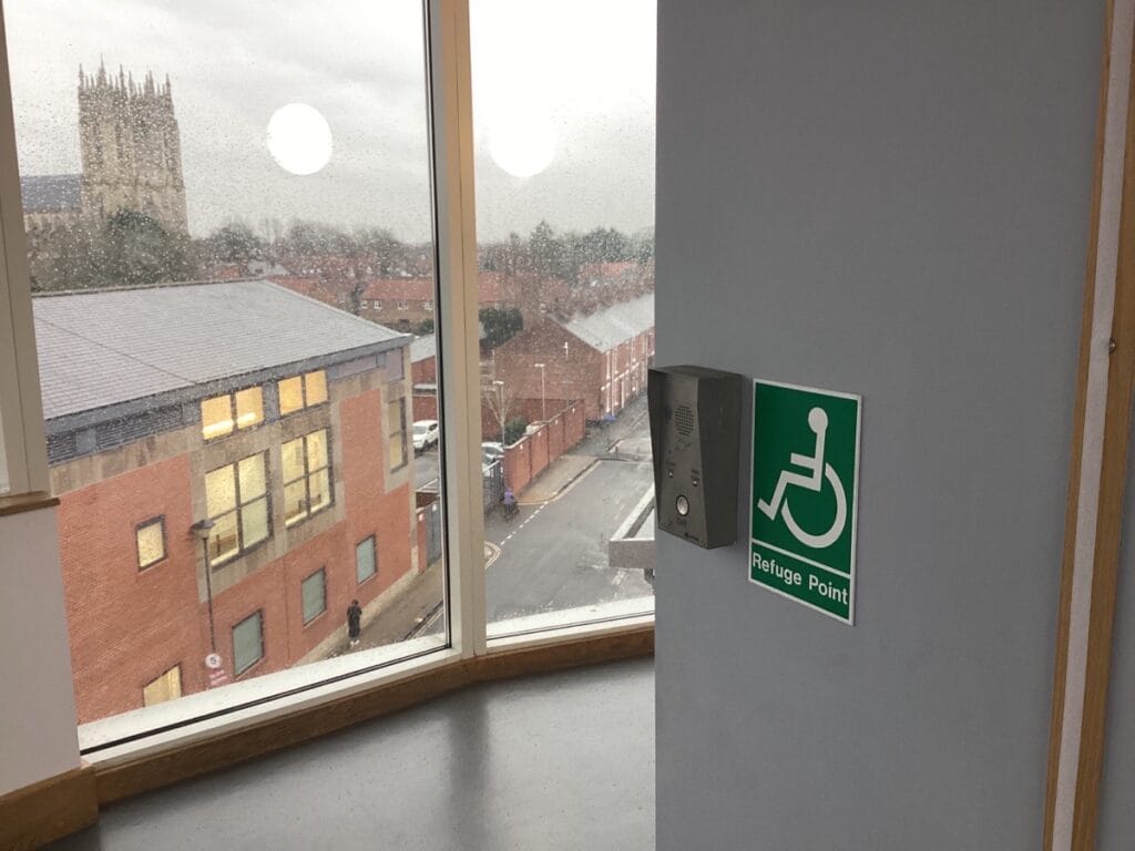 A Refuge Point sign on a grey wall with the international symbol of access printed on it next to an emergency two-way intercom within a non-specific building interior. In the background, a floor to ceiling window reveals a church and several houses being battered by the rain on a cloudy day.