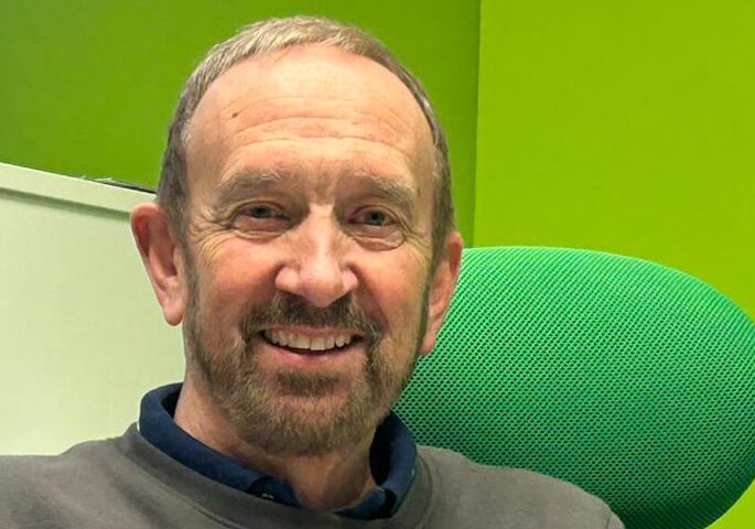 A close up of a smiling middle aged Caucasian man smiling for a photograph. He is sat in a green desk chair wearing a brown sweater.