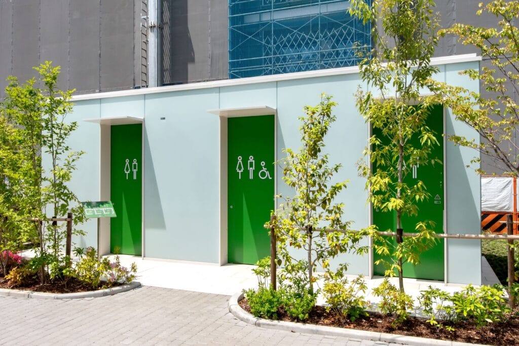 A public toilet exterior in Shibuya Ward Tokyo. Three green doors with different symbols representing genders and disability are imprinted on each. in front of the restroom, which is a small, rectangular, white building are a variety of thin trees and plants. Text overlayed on the image reads "public restrooms should be accessible to all". A public toilet exterior in Shibuya Ward Tokyo. Three green doors with different symbols representing genders and disability are imprinted on each. in front of the restroom, which is a small, rectangular, white building are a variety of thin trees and plants. Text overlayed on the image reads "public restrooms should be accessible to all".