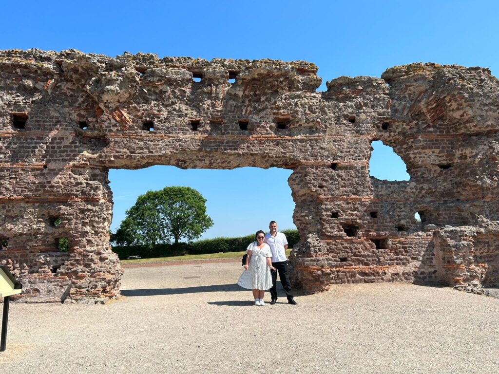 A general view of the Wroxeter Roman City ruins on a bright sunny day. The sky is a deep sky blue. Stood in front of the ruins is a short, young white woman in a white dress and sunglasses, and a slightly taller white man in a white t-shirt and black trousers.