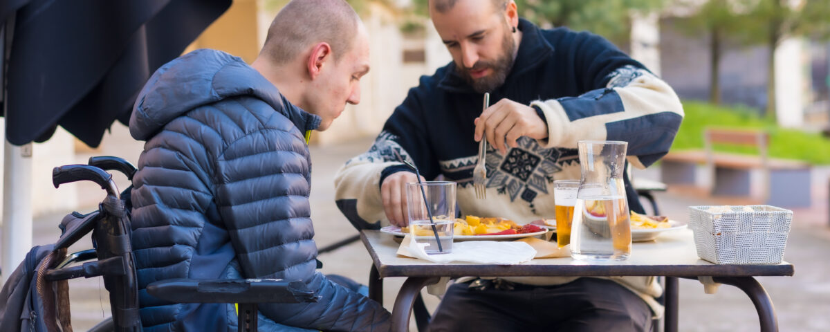 A young Caucasian man who is a wheelchair user eats on the terrace of a restaurant with a Caucasian male friend. The friend assists by cutting his food up with a knife and fork.