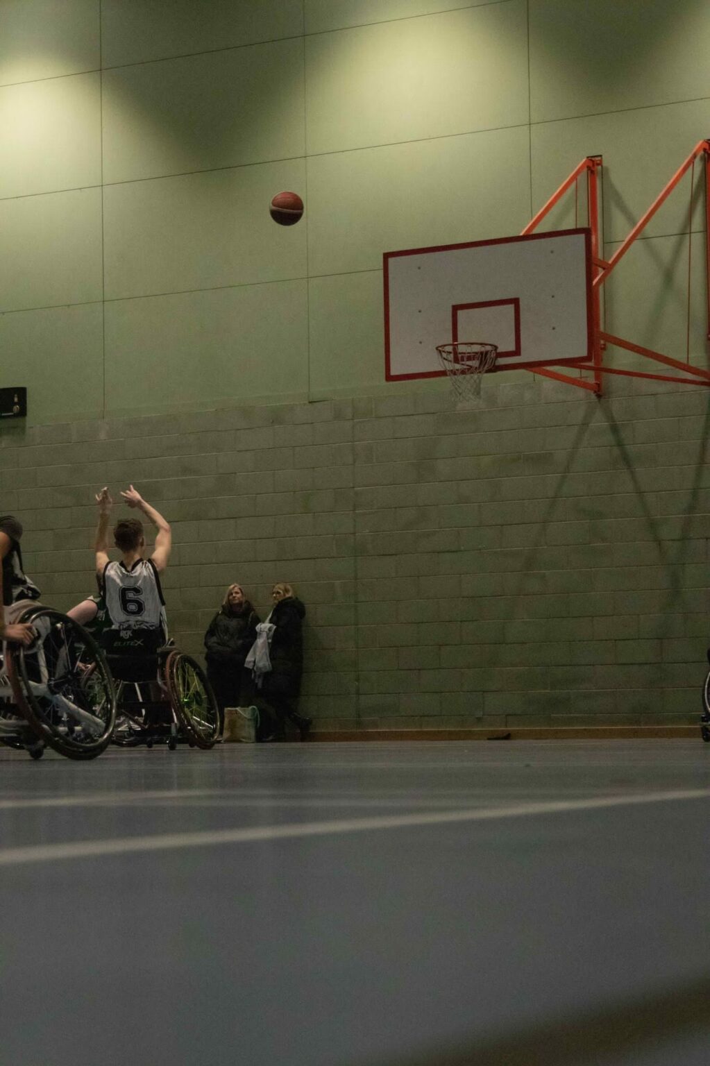 A young male Caucasian teenager in a wheelchair dressed in dark blue and white sportswear on an indoor basketball court. His arms are in the air after throwing a basketball to the hoop. The ball is mid-air.