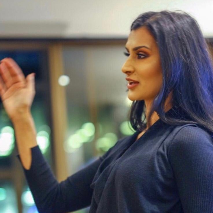 A photograph of a woman with long black hair, Safina Ahmad, gesturing her hand out while giving a speech.