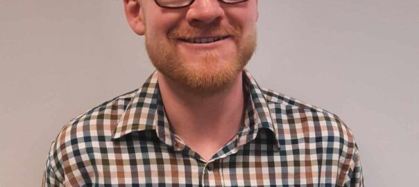 A bald Caucasian man smiles for a portrait photograph with a cream-coloured war behind him. The man has a ginger beard and is wearing black prescription glasses.