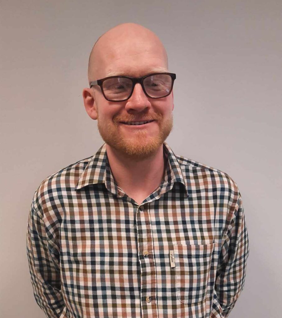 A bald Caucasian man smiles for a portrait photograph with a cream-coloured war behind him. The man has a ginger beard and is wearing black prescription glasses.