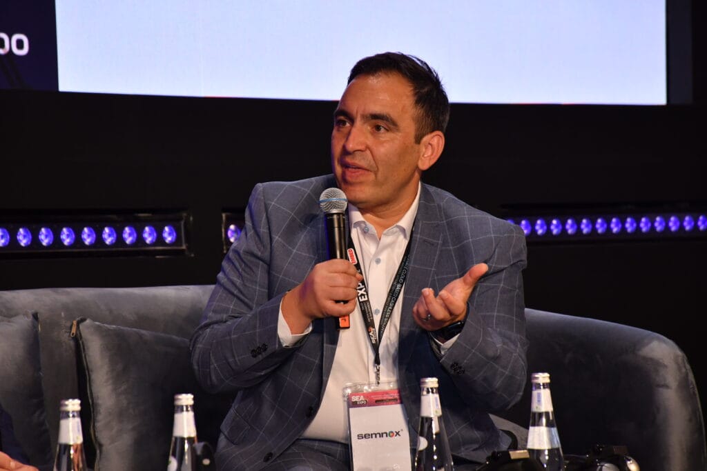 A middle aged Maltese man in a pink polo shirt and light-blue checkered suit jacket sits on a sofa in an auditorium. He is holding a microphone and holding his hand out as he says something to an off-screen interviewer.