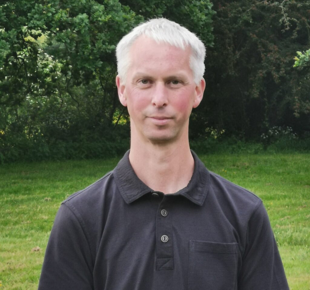 A close-up photo of a middle aged Caucasian man standing in a field wearing a black polo shirt smiles for a photograph, behind him are several trees.