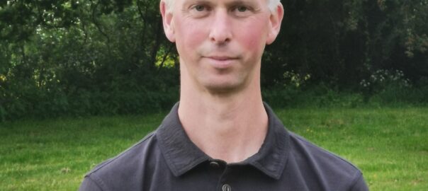 A close-up photo of a middle aged Caucasian man standing in a field wearing a black polo shirt smiles for a photograph, behind him are several trees.