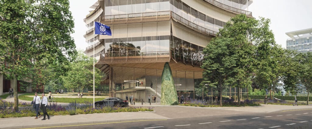 A render of the upcoming United Nations International Center for Migration, a building with large glass windows that is generally see-through. On the street front is a black car, a flagpole with a blue UN glad and people dressed in suits walking along the pavement.