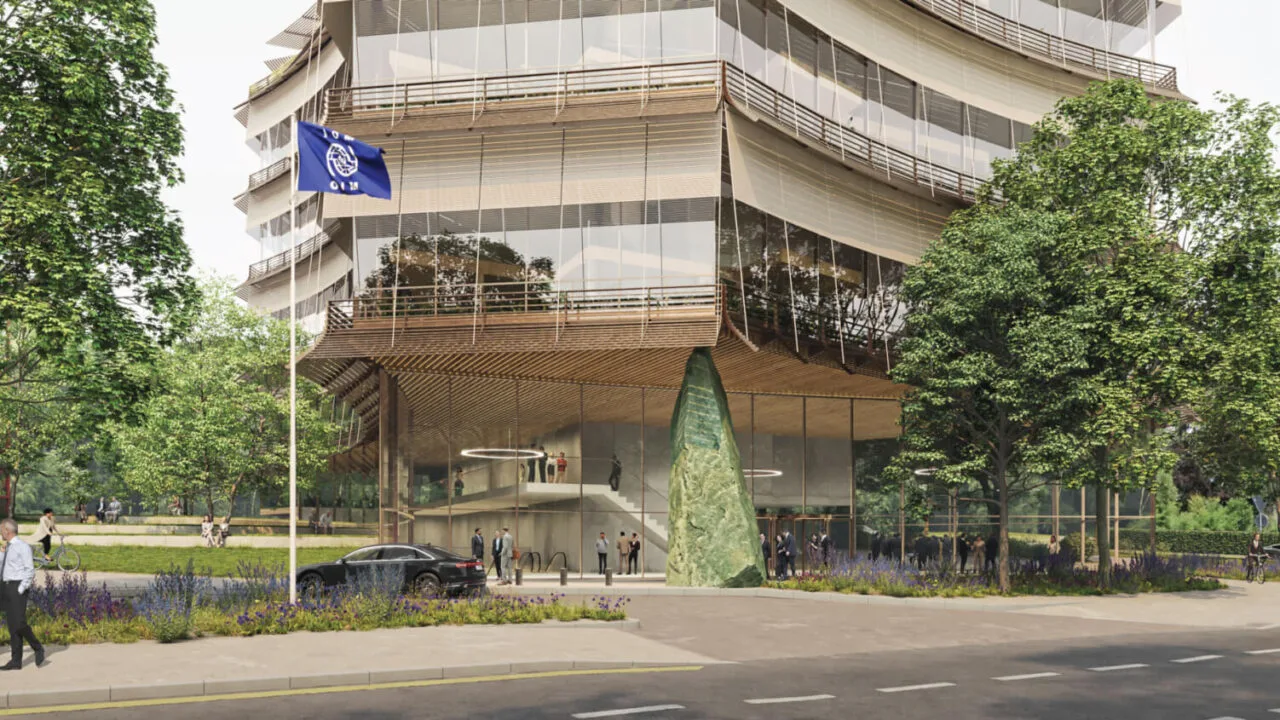A render of the upcoming United Nations International Center for Migration, a building with large glass windows that is generally see-through. On the street front is a black car, a flagpole with a blue UN glad and people dressed in suits walking along the pavement.