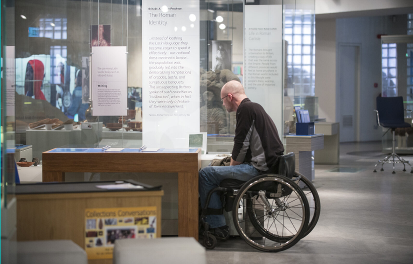 A general view of an exhibit room at the Tullie House Museum and Art Gallery populated by ancient Roman artefacts behind glass cabinets. A bald Caucasian man in a wheelchair leans forward to examine some information cards on a table.