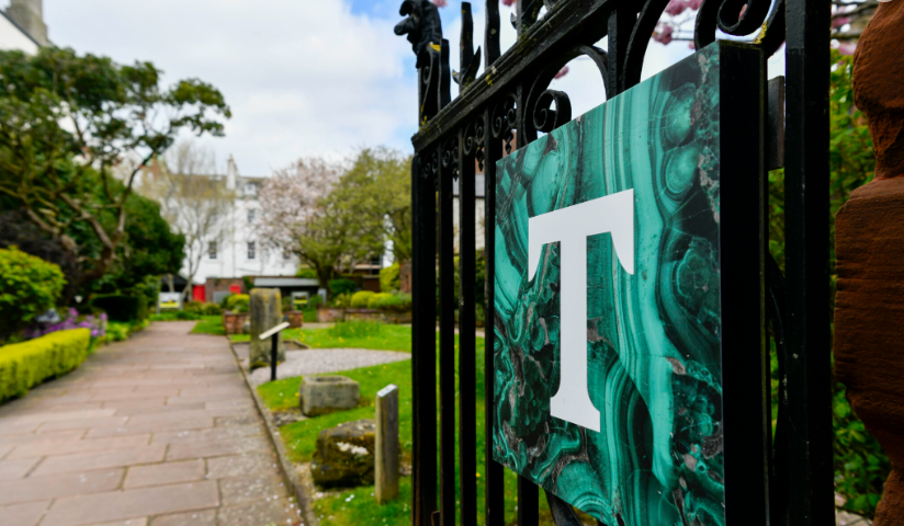 A close up photograph of an open gate leading to a walkway surrounded by a lush green garden. On the gate is a square sign displaying a white capital T, representing the Tullie Museum and Art Gallery.