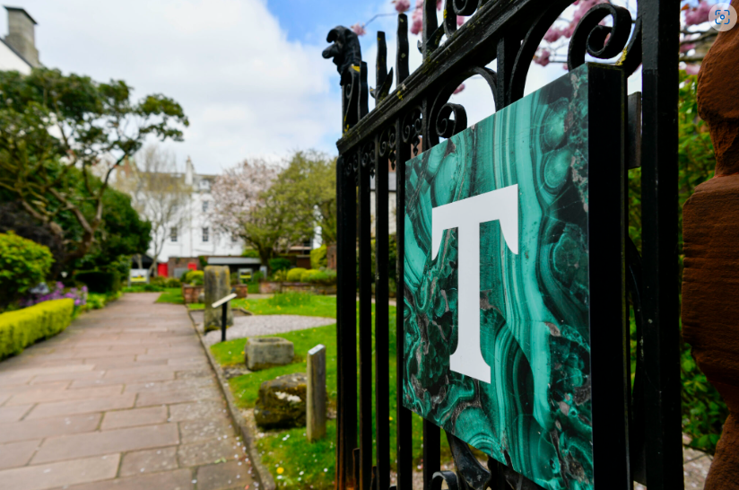 A close up photograph of an open gate leading to a walkway surrounded by a lush green garden. On the gate is a square sign displaying a white capital T, representing the Tullie Museum and Art Gallery.