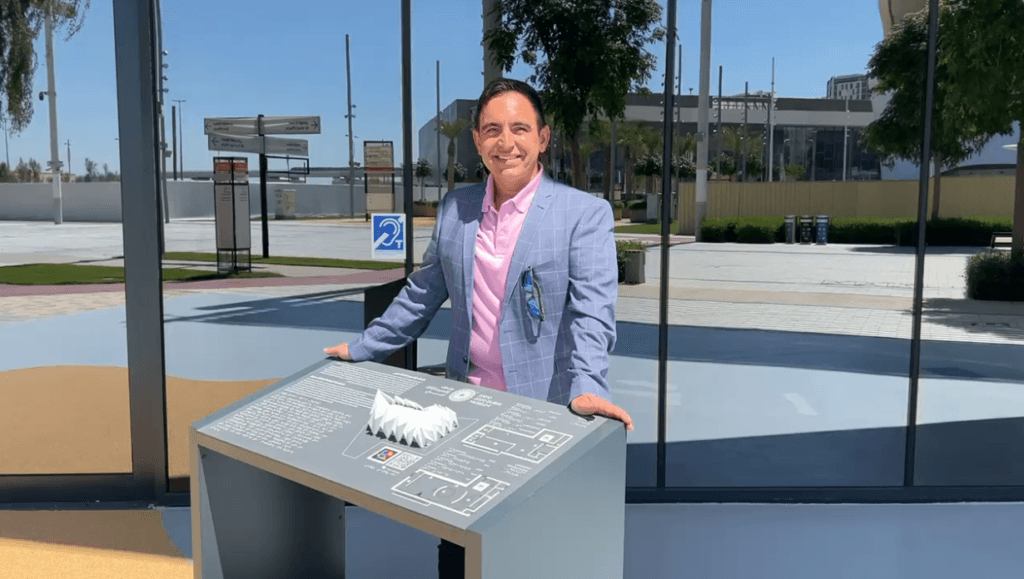A middle aged Maltese man in a pink polo shirt and light-blue checkered suit jacket standing behind a Tactile Map Board on a sunny day. His hands are placed either side of the board, which is grey and has a white miniature model of a building on it.