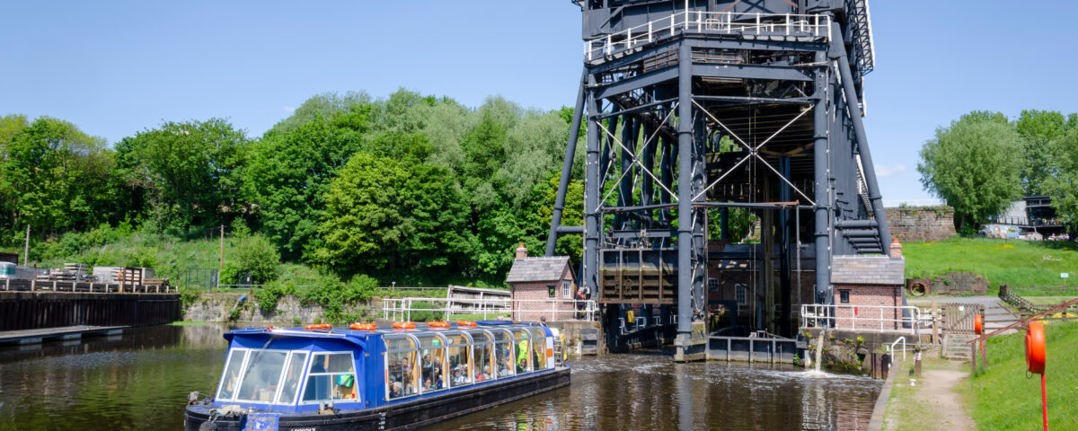 An image of the Anderton Boat Lift a mechanical structure designed to provide a 50-foot vertical link between two navigable waterways: the River Weaver and the Trent and Mersey Canal as seen from ground level. A canal boat filled with passengers makes its way along the Mersey Canal.