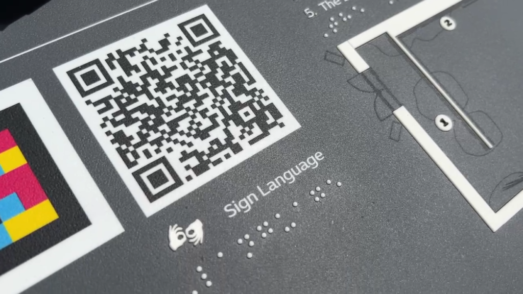 A close-up photograph of a QR code on a grey tactile map board. Underneath the QR code text reads "Sign Language" and a translation of the text in white raised braille dots.
