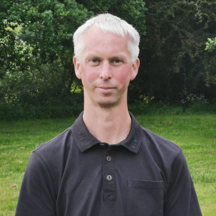 A close-up photo of a middle aged Caucasian man standing in a field wearing a black polo shirt smiles for a photograph, behind him are several trees.