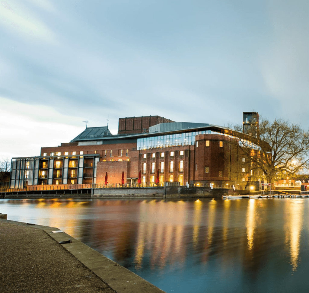 An image of a graphic showing the exterior of a large, modern red-bricked building, the Swan Theatre, during dusk. Lights surrounding the building illuminate the structure.