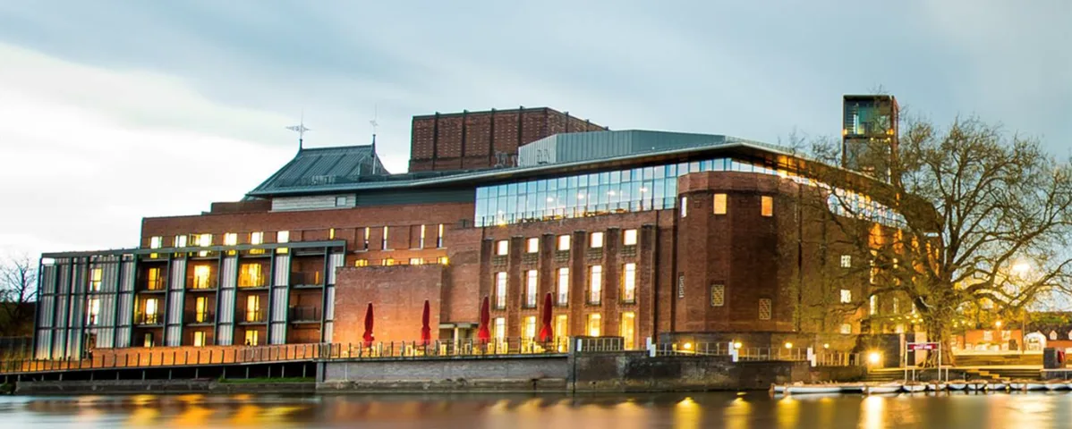 Shakespeare An image of a graphic showing the exterior of a large, modern red-bricked building, the Swan Theatre, during dusk. Lights surrounding the building illuminate the structure.