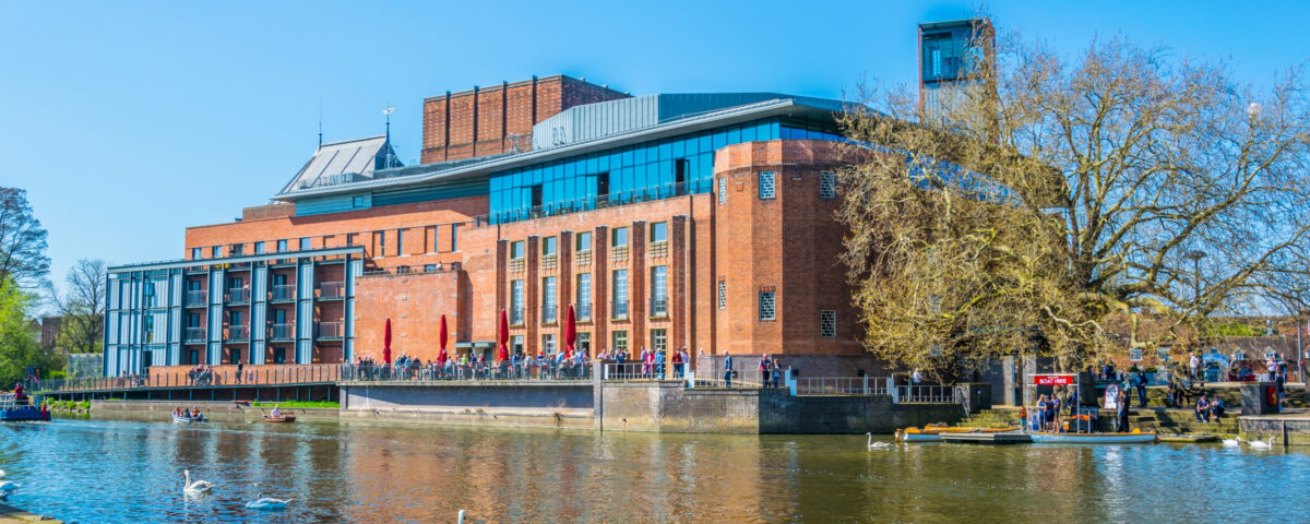 An image of showing the exterior of a large, modern red-bricked building, the Swan Theatre, on a sunny day. The building hosts the Royal Shakespeare Company. In the foreground of the image, geese can be seen paddling on the River in front of the building.