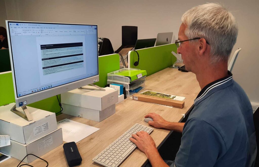 A middle aged Caucasian man with grey hair wearing a blue white polo shirt reviews a document on a Mac PC within an office environment.