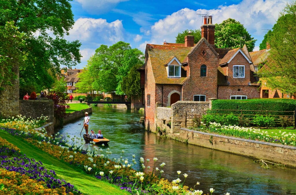 A detached, red-bricked, two-story house on a river edge in Canterbury on a bright, sunny day. The building has a beautiful garden with white flowers in front of it, across the river bank the same white flowers can be found along with purple and orange flowers. On the river, a man rows a couple on a boat across the gentle water.