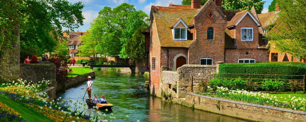 A detached, red-bricked, two-story house on a river edge in Canterbury on a bright, sunny day. The building has a beautiful garden with white flowers in front of it, across the river bank the same white flowers can be found along with purple and orange flowers. On the river, a man rows a couple on a boat across the gentle water.