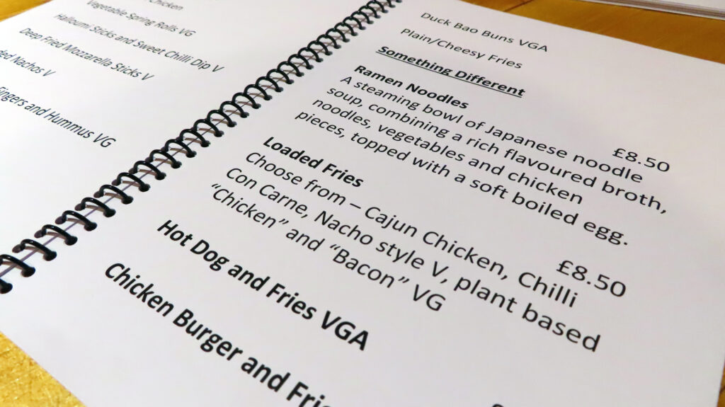 A close-up photograph of a page from the Giant Print Menu for 34 Pepper Street café showcasing different items and their descriptions. Items listed include Ramen Noodles, Loaded Fries, Hot Dog and Fries VGA, and Chicken Burger and Fries along with an eight pound fifty price tag.