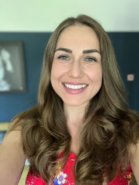 A photograph of a Caucasian woman with long brown hair smiles and poses for a selfie. She is wearing a red dress shirt with flowers in different colours on it.