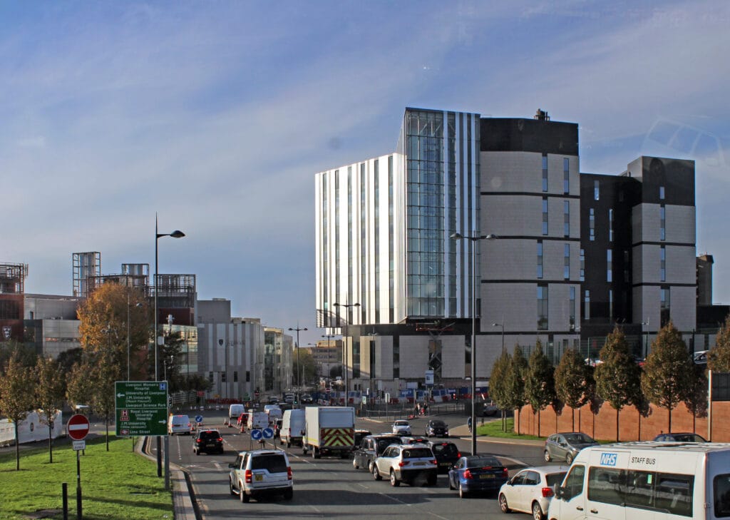 An image of the exterior of the A panoramic view of the Royal Liverpool University Hospital on an overcast day. To the left of the hospital is a busy dual carriageway with a variety of vehicles being driven on it.