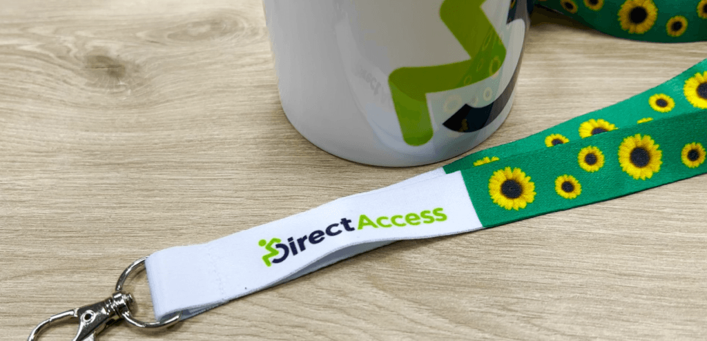 A lanyard with images of sunflowers printed on it, symbolic of people with invisible or hidden disabilities on a laminate wooden table next to a Direct Access branded mug.