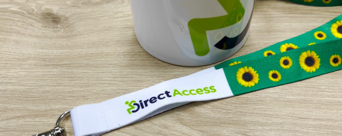 A lanyard with images of sunflowers printed on it, symbolic of people with invisible or hidden disabilities on a laminate wooden table next to a Direct Access branded mug.
