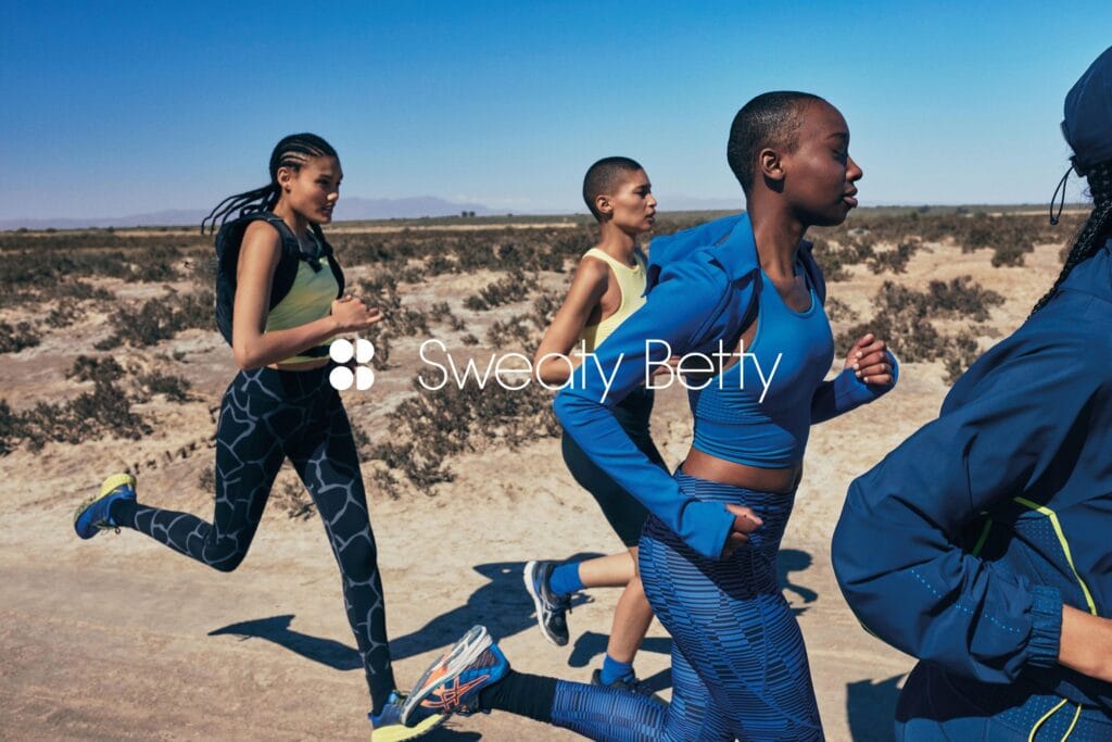 Four black women wearing vests and sweatpants running on a desert road on a sunny day. The sky is a deep blue, and in the far background are mountains.
