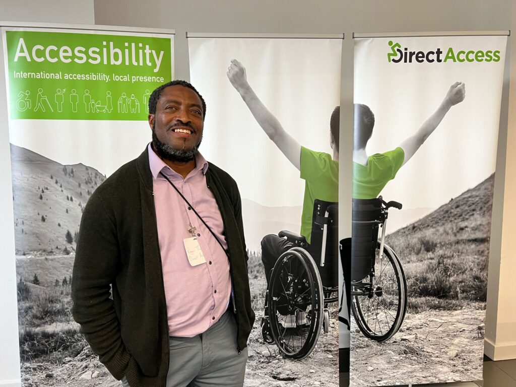 A black man wearing a pink polo shirt, grey trousers, and a black cardigan poses in front of a Direct Access exhibition banner which has a grayscale image of a young man in a wheelchair on a cliffside holding his arms in the air.