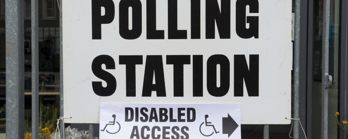 A close-up photograph of a white laminated sign hung up by string on a fence in what we would assume to be a high street. In bold, black, capitalised letters, the sign has text which reads “polling station”, with a small piece of paper sellotaped underneath it reading “disabled access”, also in bold, black, capitalised letters.
