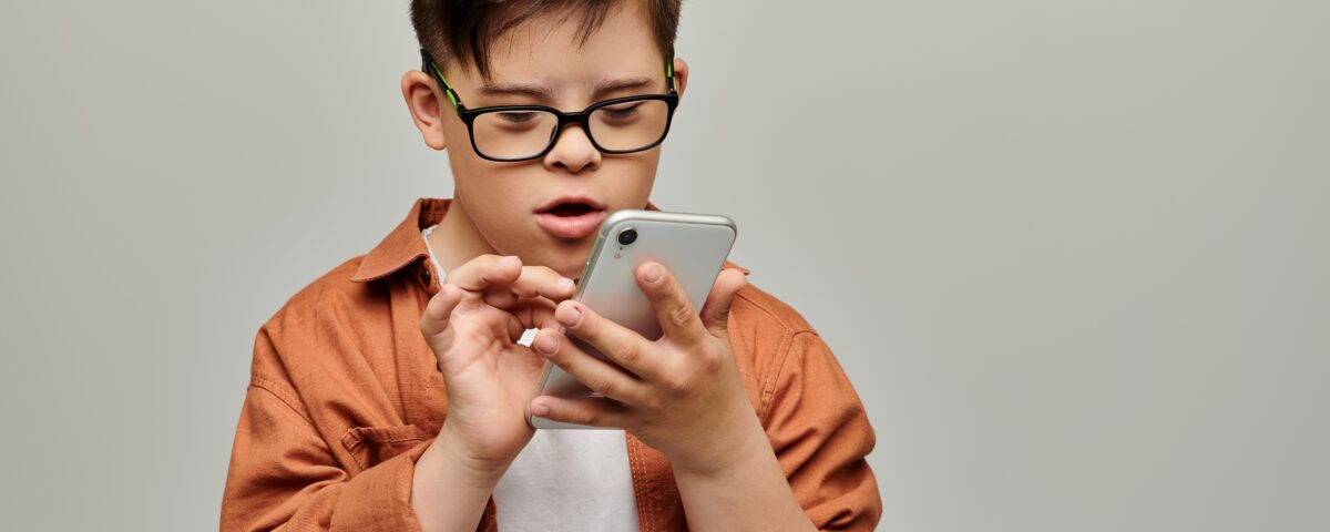 A little boy with down syndrome wearing glasses focuses intently on a mobile phone, scrolling on the screen.