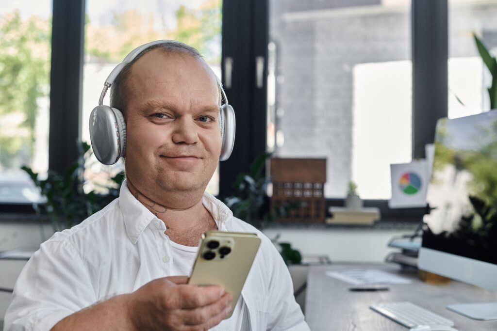 A smiling Caucasian man wearing silver coloured headphones holds his phone within an office environment.