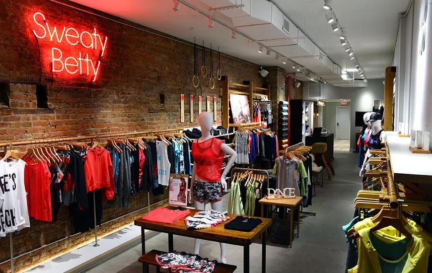 The interior store floor of a Sweaty Betty; a womenswear retailer. Mannequins modelling clothes are displayed in the center of the room. On the left wall is a glowing red LED Light fixture which spells Sweaty Betty. Directly underneath is a long row of clothes displayed on racks.