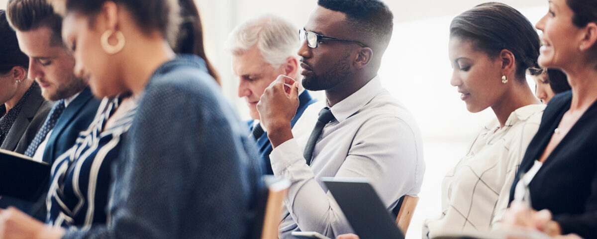 Shot of a mixed group of businesspeople sat on different rows of seats attending a class. Some are watching an out of shot person attentively, while others are writing information on their notebooks, laptops, and tablets.