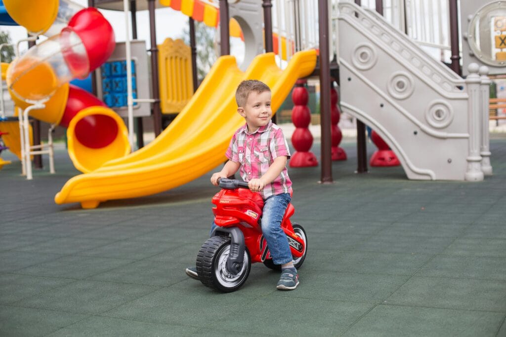 A young caucasian boy (around 6 or 7) on a plastic motorbike smiles and looks into the distance, presumably at a parent, in a children's playground. Behind him are slides, tube slides and other interactive playground features.