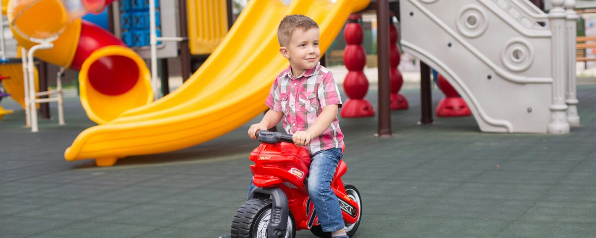 A young caucasian boy (around 6 or 7) on a plastic motorbike smiles and looks into the distance, presumably at a parent, in a children's playground. Behind him are slides, tube slides and other interactive playground features.