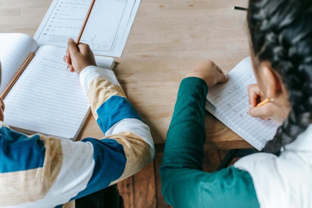 A male and female student sitting next to each other in a classroom behind a brown wooden desk write in their notebooks.