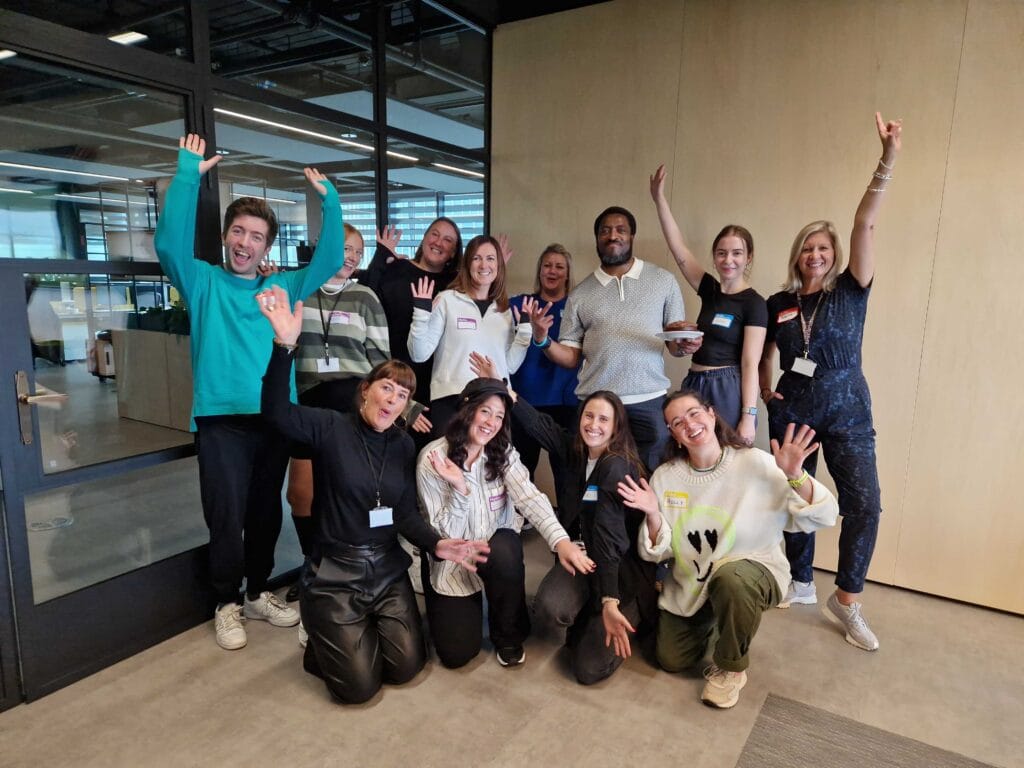 A mixed group of adults pose for a group photograph at the end of their accessibility awareness training in the corner of a boardroom. All are smiling and some people are thrusting their arms in the air energetically. A few adults are kneeling on the floor.