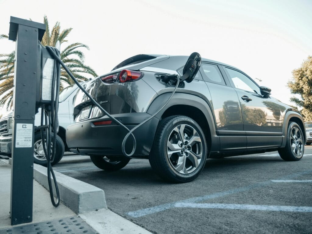 A black electric vehicle as seen from behind with its fuel cap open. The vehicle is plugged into an Electric Vehicle charging point in a parking lot. Behind the car are palm trees.