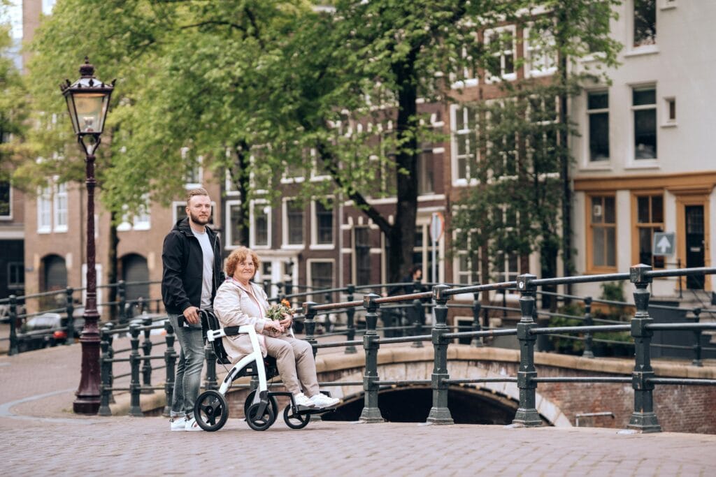 An elderly Caucasian woman in a stroller holding flowers is being pushed by a younger man, presumably a son or grandson. They are walking along the canal side in Amsterdam.