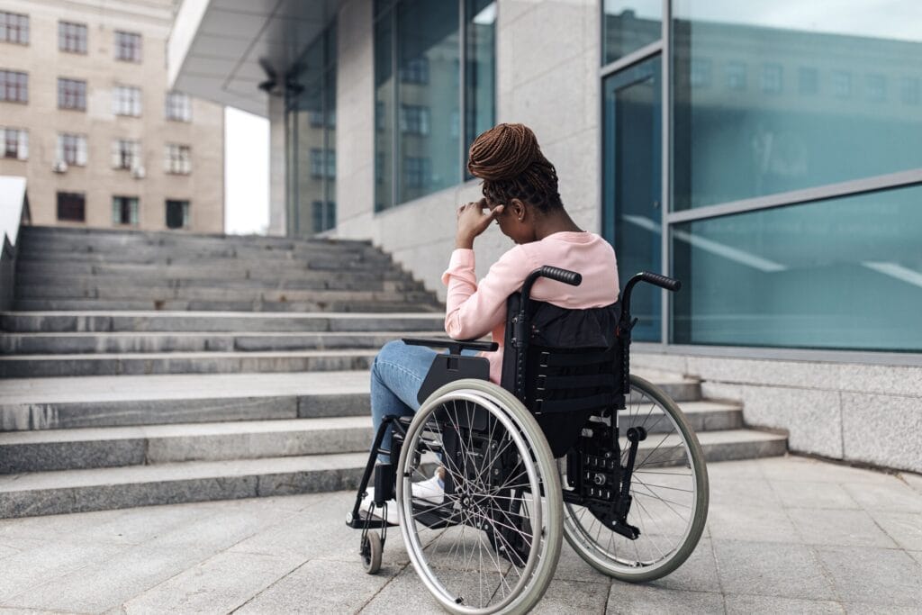 An unhappy young Black Woman in a wheelchair looking fed up in front of a staircase with no ramp access. She is holding her head in her hand.